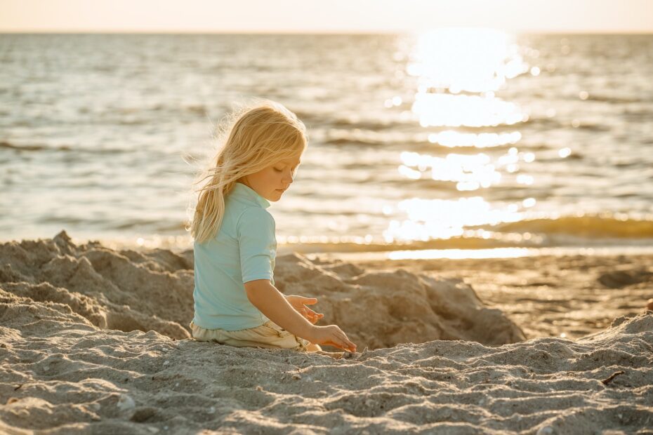 blond girl sitting on a sandy beach and sun reflecting in the sea
